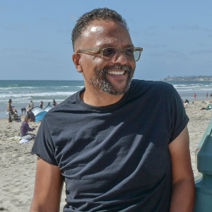 David Odusanya wearing a black t-shirt, standing against a lamppost in front of a beach