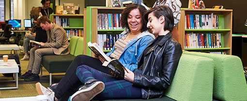 Photograph of two students sitting together in a library, each reading their own book.