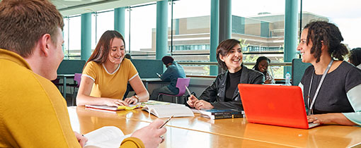 Students working around a large table, using laptops and notepads as they converse