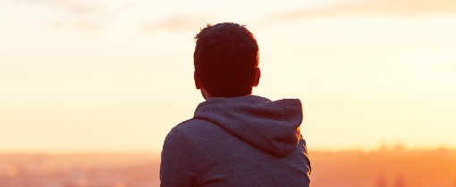 A person wearing a grey hoodie looking into a hazy orange sunset.