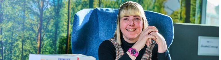 Female student sitting at table in Education building with books in front of her smiling at camera
