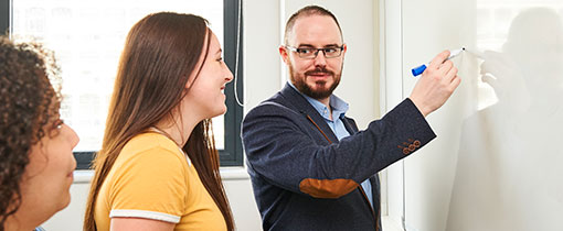 A lecturer writing on a whiteboard, turning to face two students who are watching him