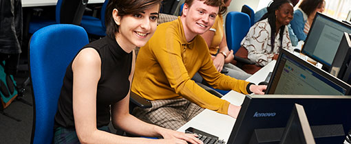 Photograph of students sitting at computer desks in blue office chairs, looking at monitors and typing on keyboards.