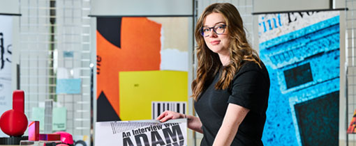 An art student with long brown hair and glasses stands in a creative workspace, holding a large printed design featuring bold text. She wears a black dress and looks towards the camera with a confident expression. The background includes colourful graphic design posters and various creative materials.